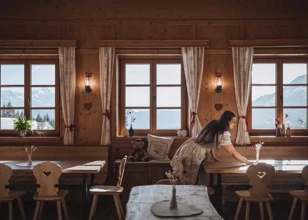 Ihre Almhütte in Tirol Frau räumt Holztisch in gemütlichem Berghotel mit Fensterblick auf Berge