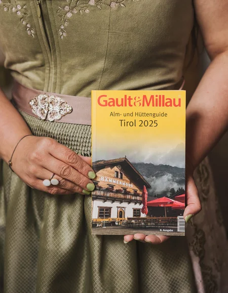 Ihre Almhütte in Tirol Frau in grünem Dirndl hält den Gault&Millau Alm- und Hüttenguide Tirol 2025