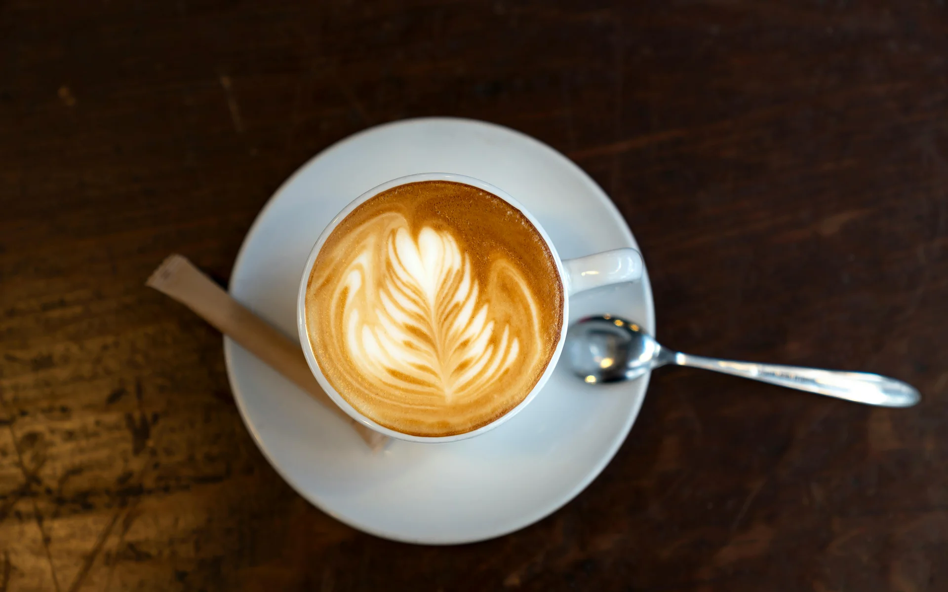 Werfen Sie einen Blick in die Bildergalerie Weiße Tasse mit Latte Art auf Holztisch mit Löffel und Zuckerstange