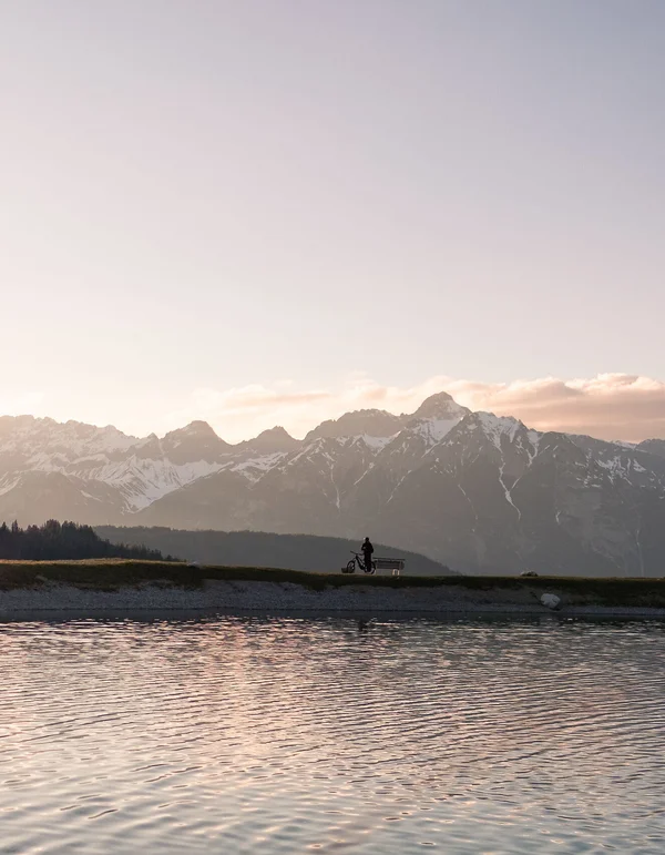 Erlebniswelt Alpenbad Leutasch Person mit Fahrrad am See vor schneebedeckten Bergen bei Sonnenuntergang