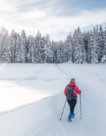 Urlaub in den Bergen: Ihr Naturhotel in Tirol Person wandert mit Stöcken auf verschneitem Weg im Winterwald bei Sonnenschein