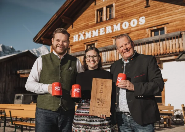 Ihre Almhütte in Tirol Drei Personen vor Holzhaus mit Bierkrügen und Auszeichnung BierWirt 2023