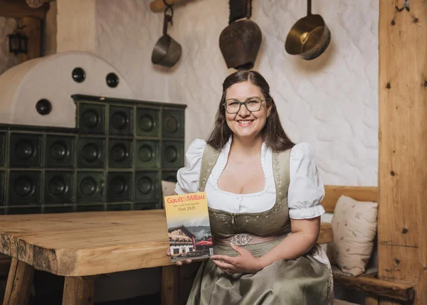 Ihre Almhütte in Tirol Frau in Tracht hält Gault&Millau Tirol 2025 Magazin in rustikalem Zimmer