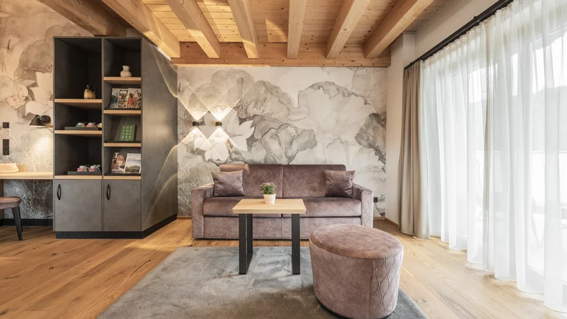 Biohotel Leutascherhof – Ihr Kraftort in den Tiroler Alpen Gemütliches Wohnzimmer mit Holzdecke, Sofa, Hocker und großem Fenster mit Vorhängen